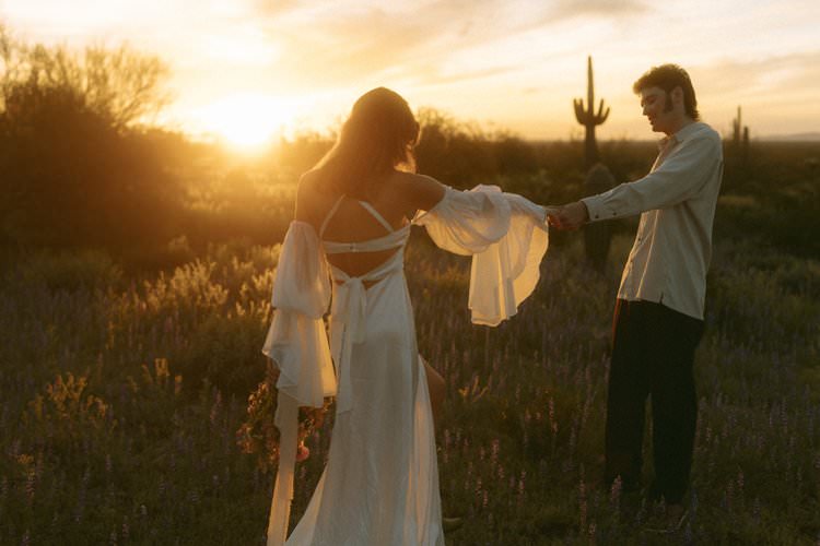 Against the backdrop of a setting sun casting a warm golden light, a couple stands hand in hand in a field of purple wildflowers. The woman, seen from the back, is wearing a flowing white dress with off-the-shoulder sleeves and a delicate tie at the back, her hair cascading down her shoulders. The man, facing the woman, is in a casual white shirt, his posture relaxed. The scene is peaceful and romantic, with silhouettes of cacti in the distance adding to the serene desert landscape.