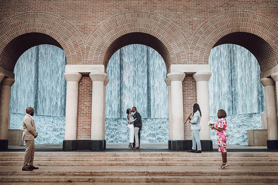 A wedding couple is kissing each other underneath one of three arches of a architectural building and their groomsman and bridesmaid is standing on steps in Houston.