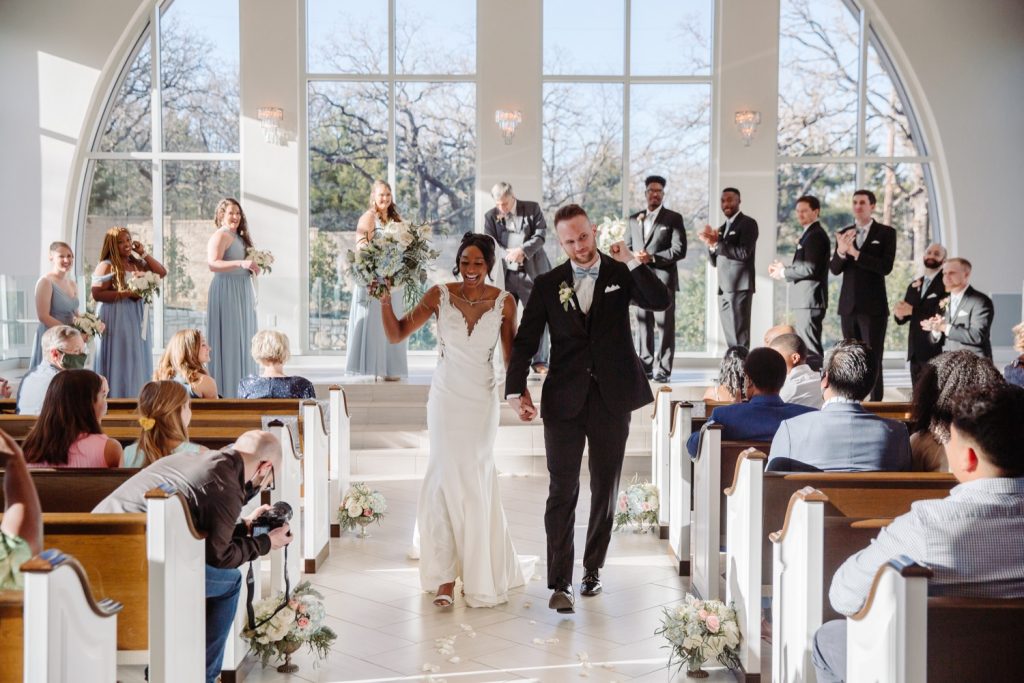 A wedding couple is walking along the aisle and is waving at their guests at the Bowden Holly Chapel, one of the wedding venues Fort Worth offers.