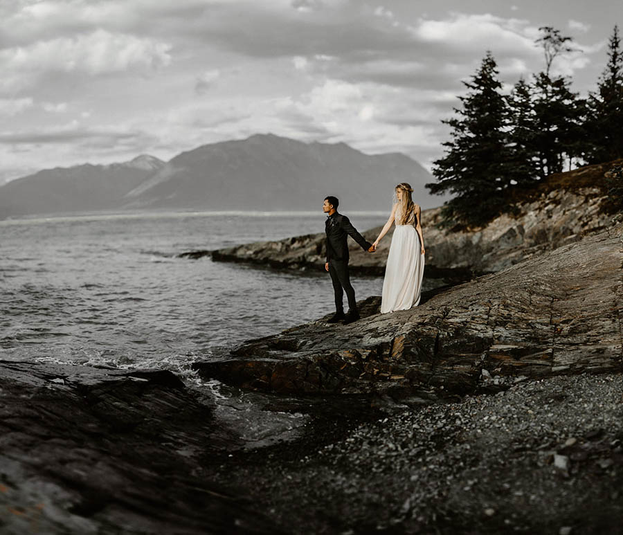 A wedding couple is standing next to the ocean on rocks and holding each others hand in Hope, Alaska.