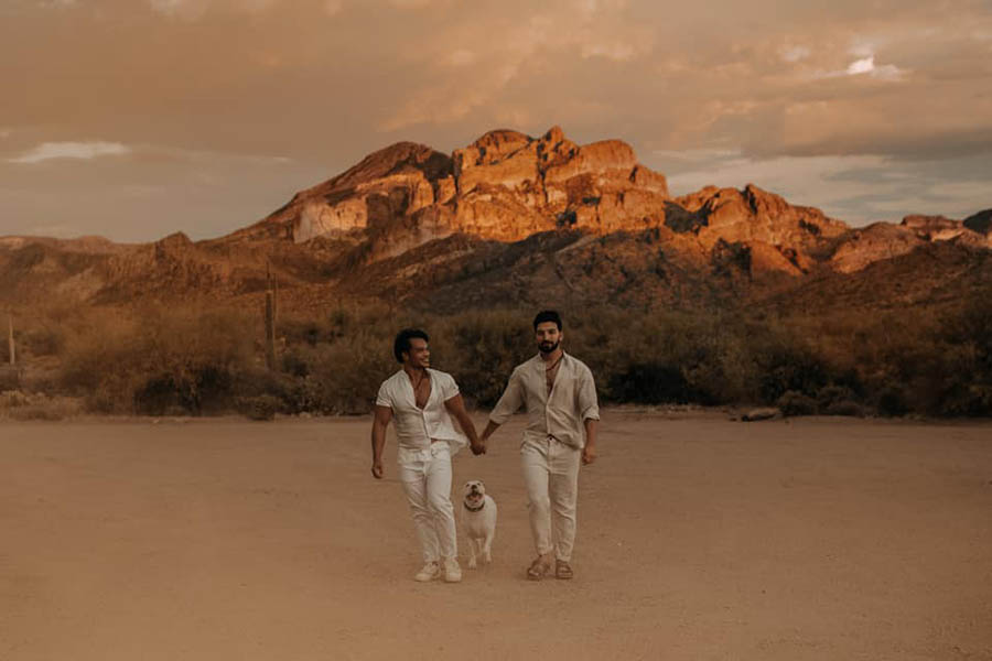 A wedding couple is holding hands and walking on a dusty area Iwrith a white dog in-between them in front of the Superstition Mountains in Arizona.