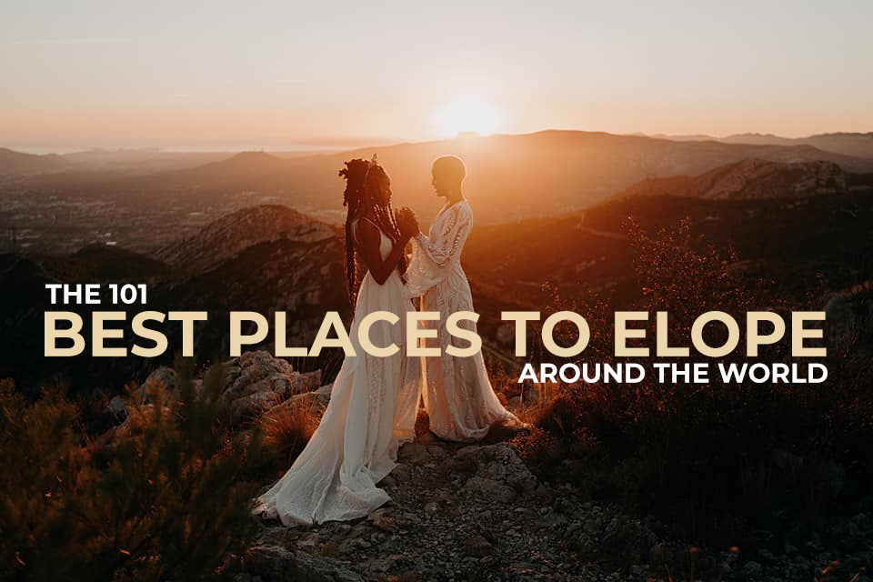 A wedding couple is standing on top of a mountain and holding hands in front of a sunset.