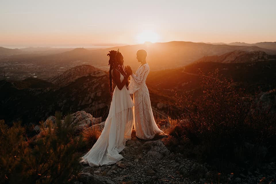 Two brides are holding hands and standing on a mountaintop with a sunset in the background.