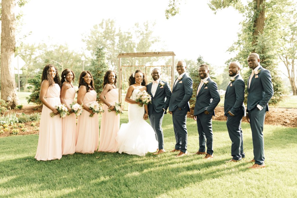 A wedding couple and their bridesmaids and gromosmen are standing outside on the lawn of the Leopold's Mississippi Gardens, one of the wedding venues Minneapolis offers.