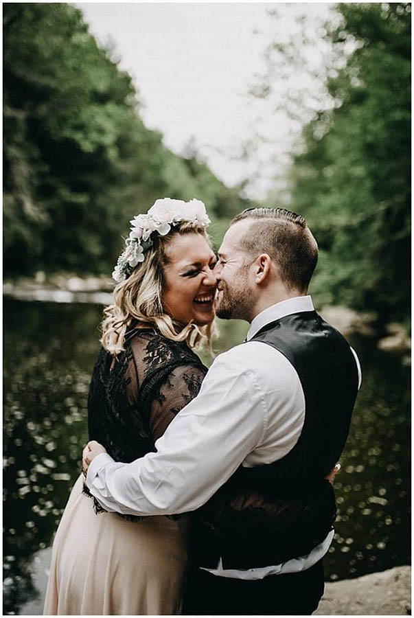 A wedding couple is holding each other and smiling at each other whilst standing in front of a lake in the Pocono Mountains. 