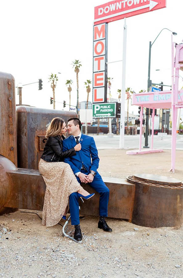 A couple is sitting on a rusty sculpture and in the background are several road signs, palmy and traffic lights, located in Las Vegas.