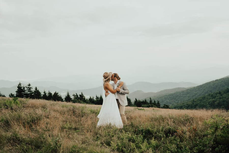 A wedding couple is kissing each other and standing in a field at the Great Smokey Mountains.