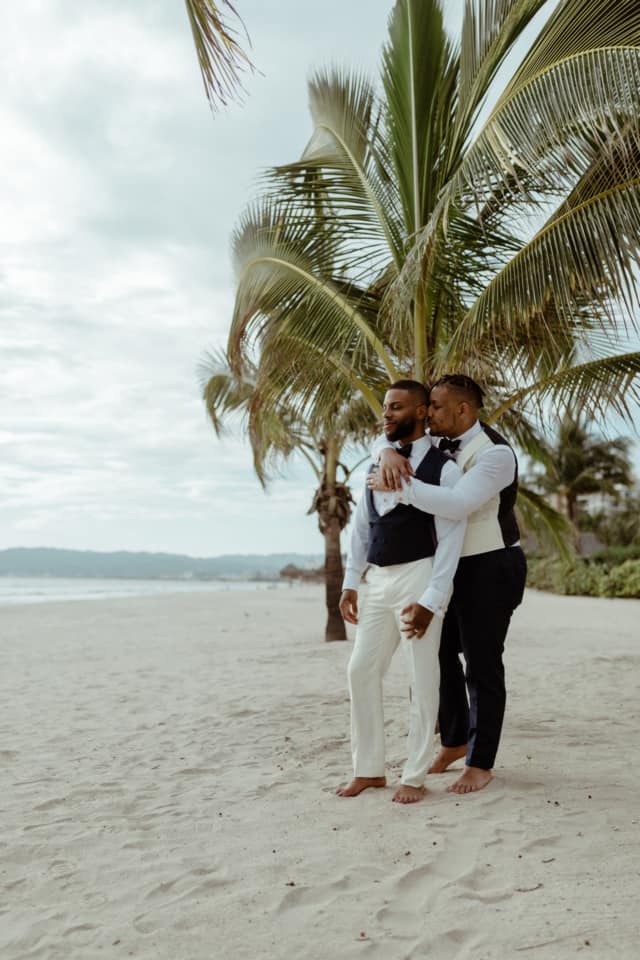 Two grooms are holding each other and standing on a beach at Puerto Vallarta.