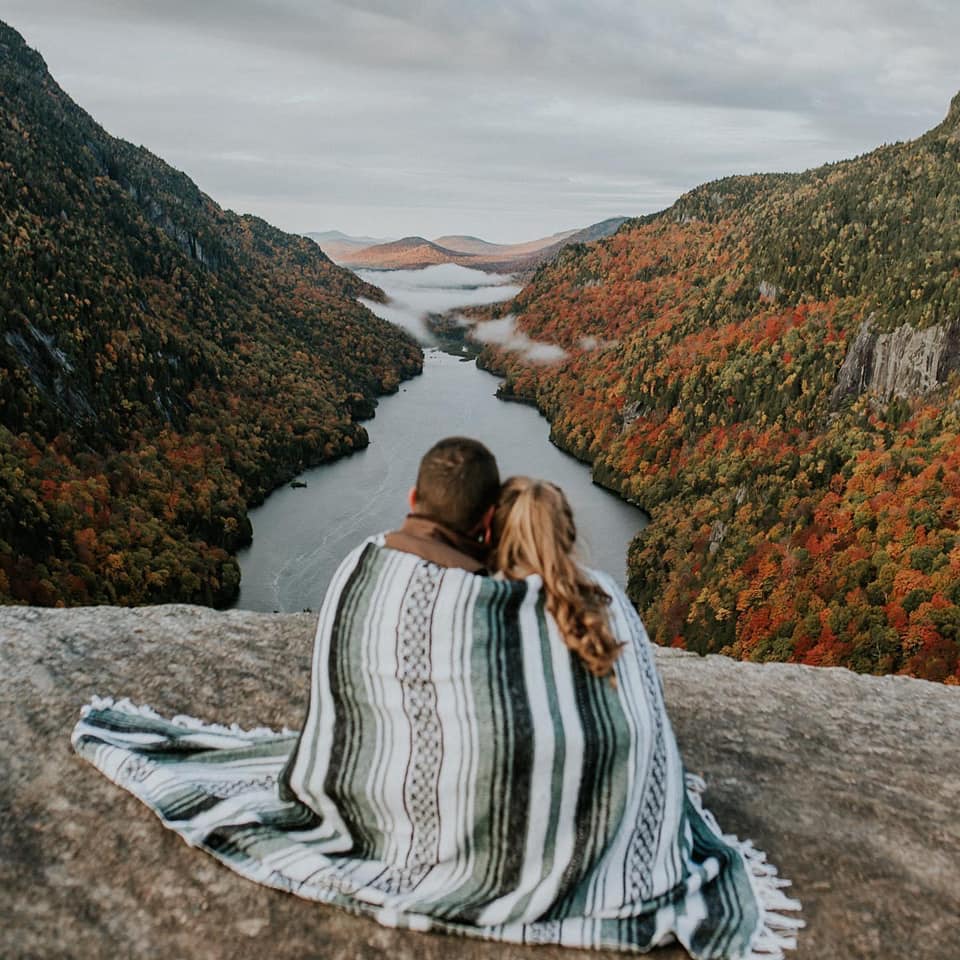 A couple is cuddling in a blanket and sitting on top of a mountain and looking to a lake in the valley at the Indianhead Mountain in the Adirondacks.