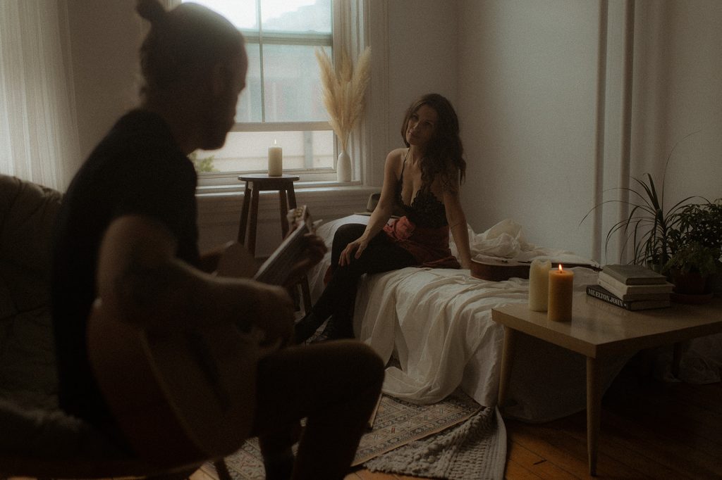 Jennifer Moher is sitting on a bed and a man is playing the guitar.