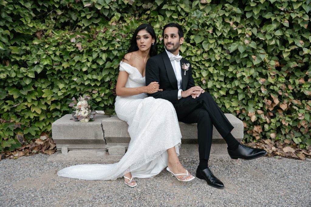 A newlywed couple poses on a stone bench in front of a lush, green ivy wall. The bride, wearing an elegant off-shoulder white gown with a long train and intricate henna designs on her hands, sits closely beside the groom, who is dressed in a classic black tuxedo with a white bow tie. A bouquet of flowers rests on the bench beside them, enhancing the romantic ambiance of wedding photography styles.