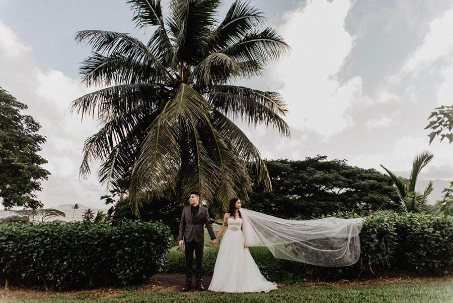 A wedding couple is holding hands and looking away from each other in the opposite direction and they are standing in front of a garden in Oahu.