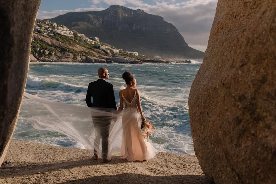 A wedding couple is standing on rocks near the ocean and is holding hands near Capetown.