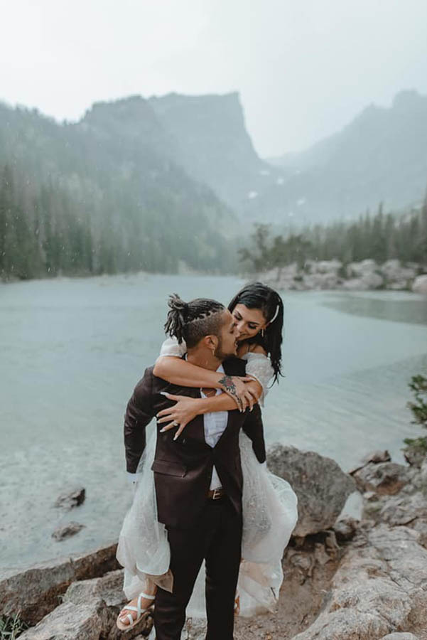 A groom is piggybacking his wife and standing on rocks, behind them is a lake that is located in the Rocky Mountains.