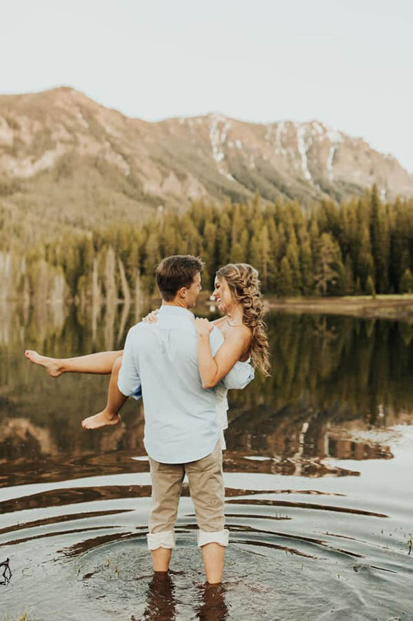 A groom is carrying his wife and standing in a lake, in the background are trees and mountains which are located near Bozeman.