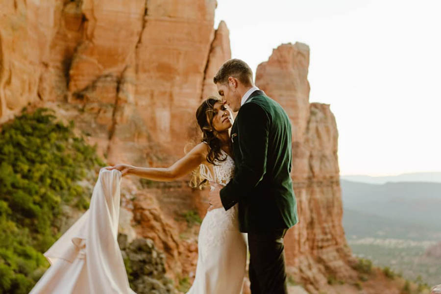 A wedding couple is holding each other and the bride is holding her dress and they are standing near mountains in Sedona.