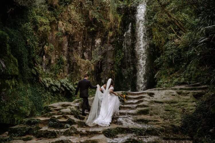 A wedding couple is walking up stairs made of rocks towards a waterfall.