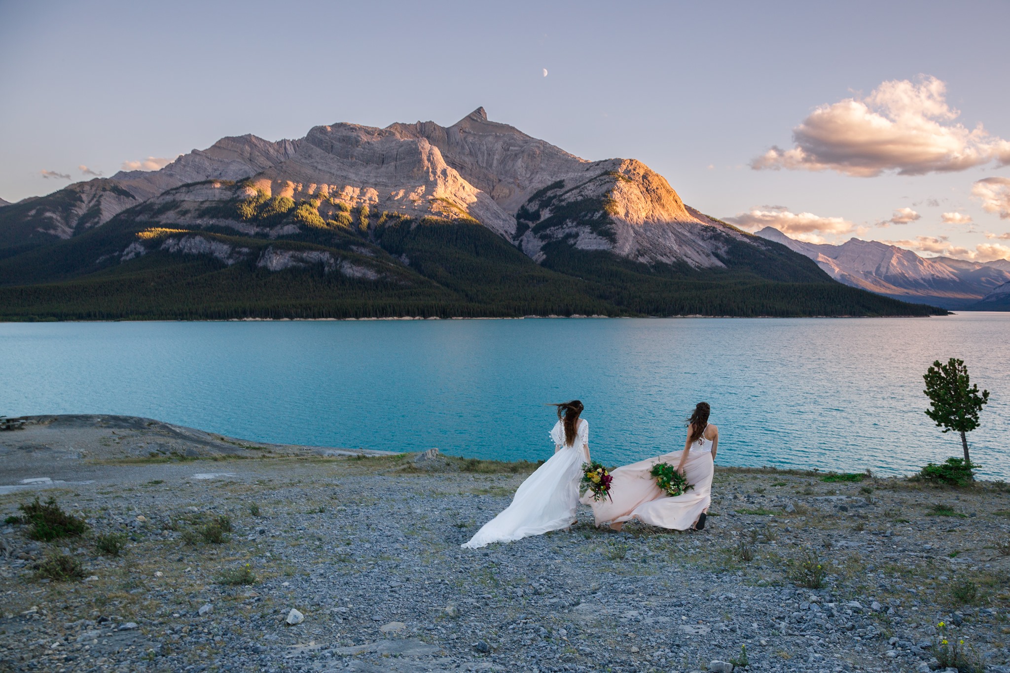 Two brides are standing in front of Abraham Lake and looking towards the lake.