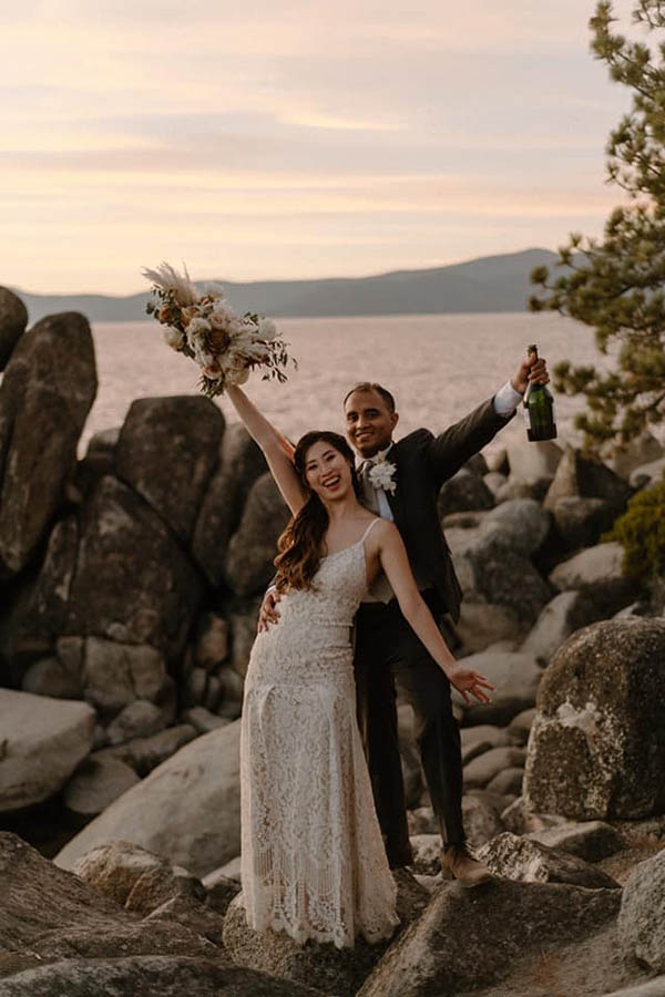 A wedding couple is standing on rocks in front of Lake Tahoe in Nevada and cheering whilst holding a bouquet and a bottle of wine.