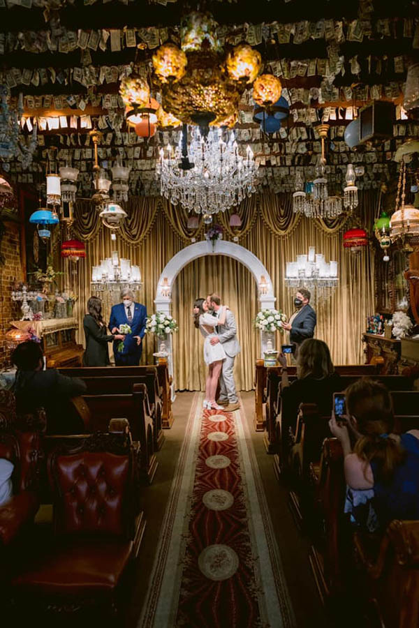 A wedding couple is kissing each other and standing in a colorful room at the end of an isle with a few wedding guests sitting next to the isle in New Orleans.