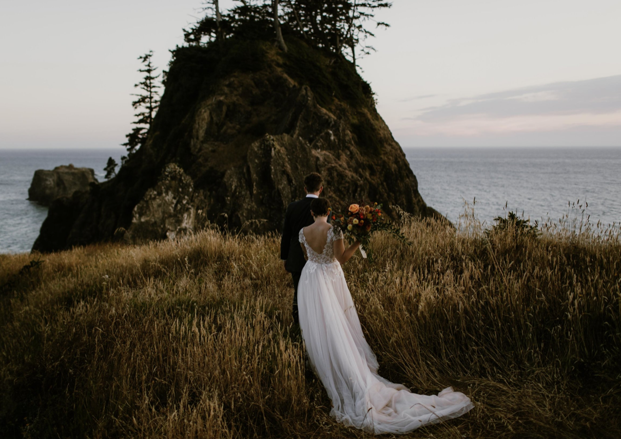 A wedding couple is walking towards a mountain with the ocean in the background. 