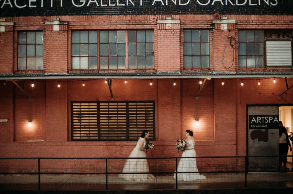 A wedding couple is standing in front of a brick wall at the LGBTQ friendly Artspace 111 Gallery and Gardens, one of the wedding venues in Texas.