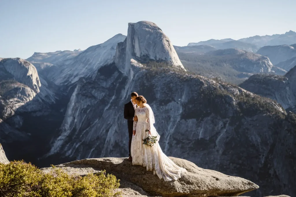 Bride and groom standing on a cliff during a Yosemite elopement with stunning views of Half Dome and mountain ranges in the background.