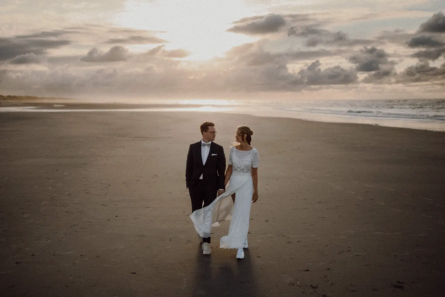 Bride and groom walking along an empty beach at sunset, with soft clouds and golden light reflecting on the sand.