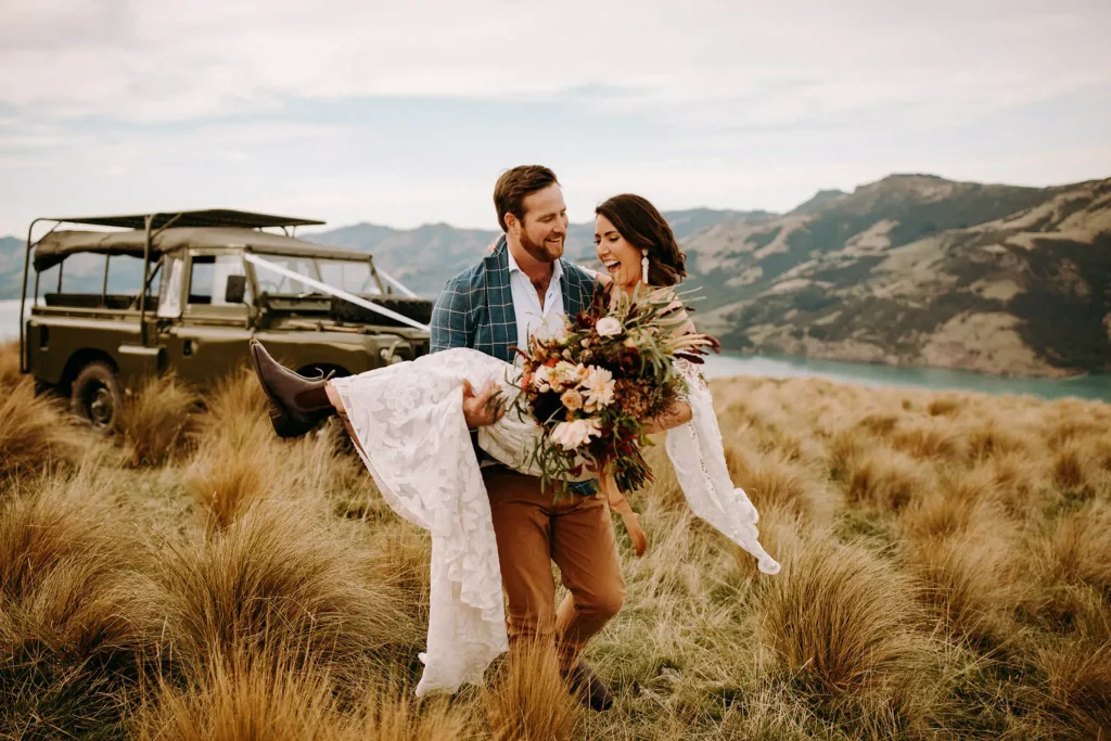 The groom carries the bride in his arms while walking on top of a hill in New Zealand. A picture part of the Checklist for destination wedding article.