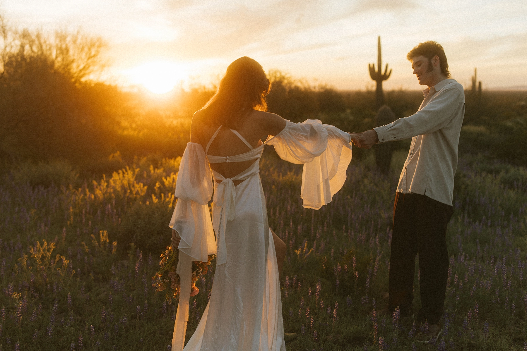 A couple holding hands in a desert field at sunset, with a woman in a flowing white dress adorned with flowers and a man in a casual white shirt, surrounded by saguaros and vibrant purple wildflowers, capturing the essence of film-like Lightroom Presets natural and romantic photography style.