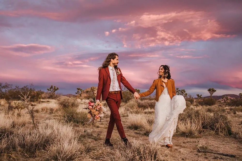 A couple walking hand-in-hand at sunset in Joshua Tree National Park, showcasing an all-inclusive wedding packages. The bride wears a white gown with a brown jacket, while the groom sports a burgundy suit, holding a bouquet of flowers.