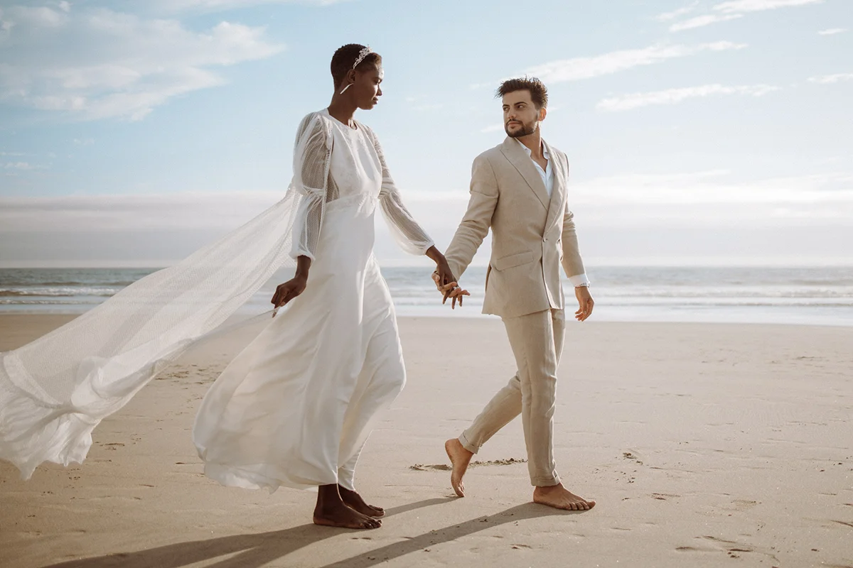 An elegant couple holds hands while walking along a sandy beach. The woman, dressed in a flowing white wedding gown with sheer sleeves and a long veil caught in the sea breeze, strides confidently beside the man. The man is in a stylish beige suit that matches the natural tones of the beach, looking at her with a serene expression. Both are barefoot, with the ocean's gentle waves in the background, symbolizing a peaceful and romantic moment shared between them.