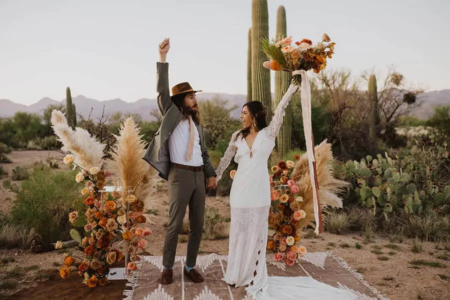 A couple is celebrating their marriage at one of the awesome elopement venues in Arizona, surrounded by cacti and desert scenery. The bride wears a lace dress and holds a bouquet, while the groom wears a suit and hat.