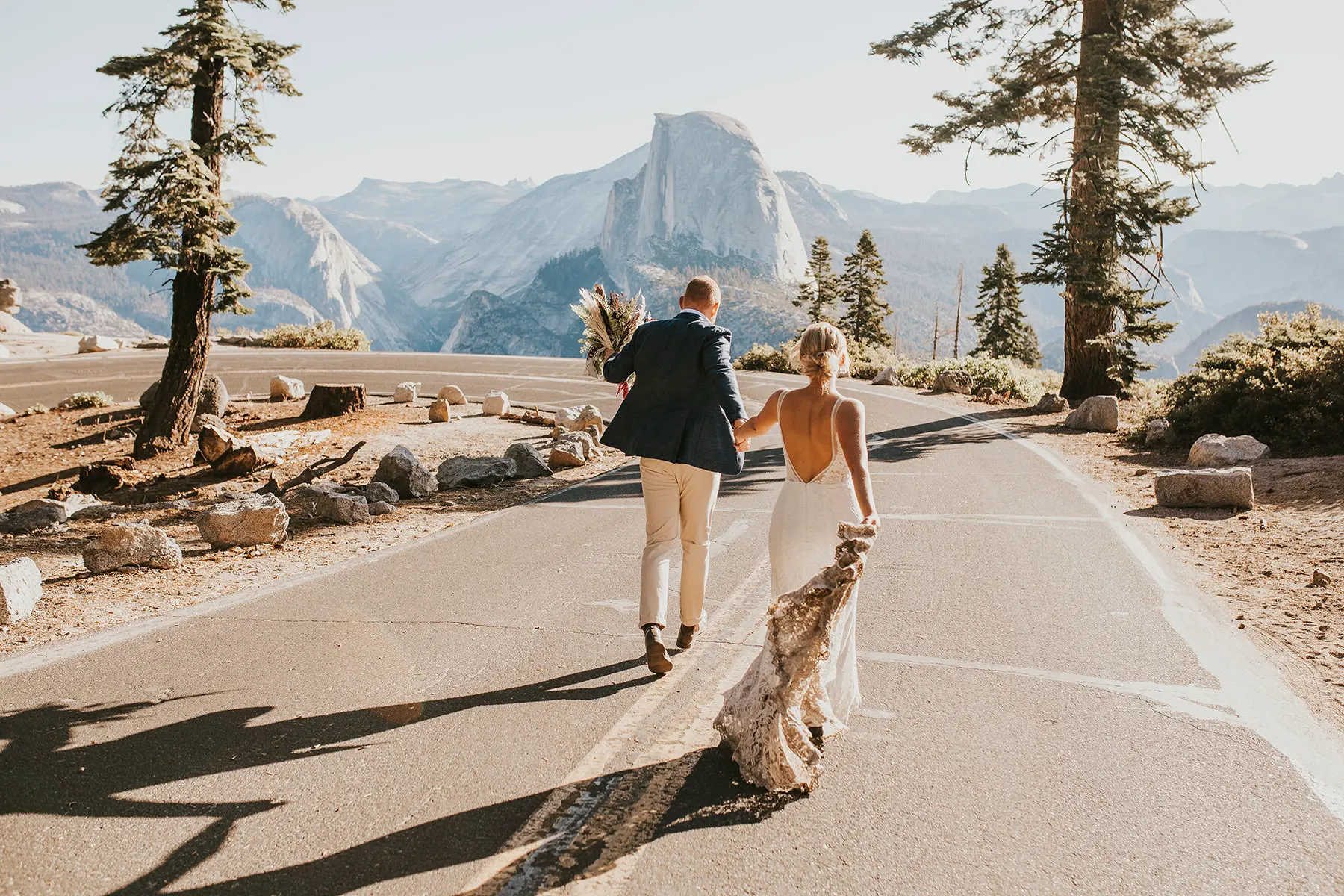 Bride and groom walking hand in hand on a winding road in Yosemite, with Half Dome and towering pines in the background, captured with one of the best lens for wedding photography.