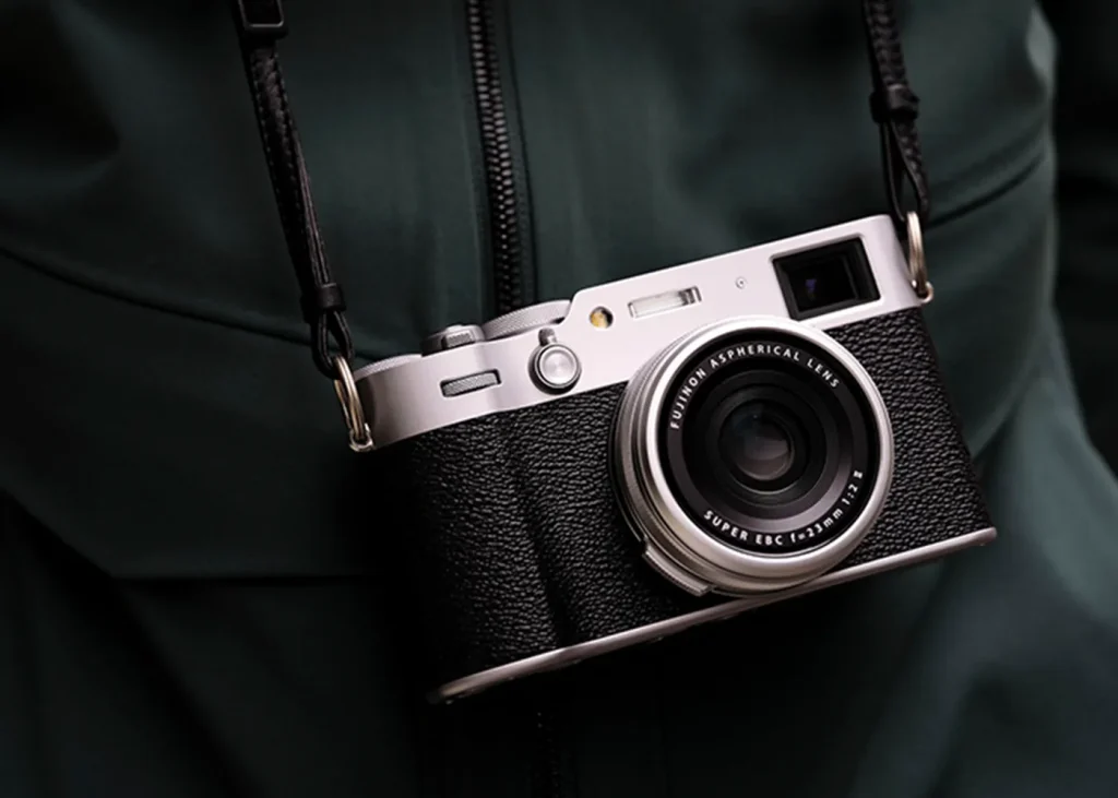 A Fujifilm X100VI with a prime lens is suspended by a black leather strap against a dark green, textured backdrop, highlighting the sleek design of the camera.