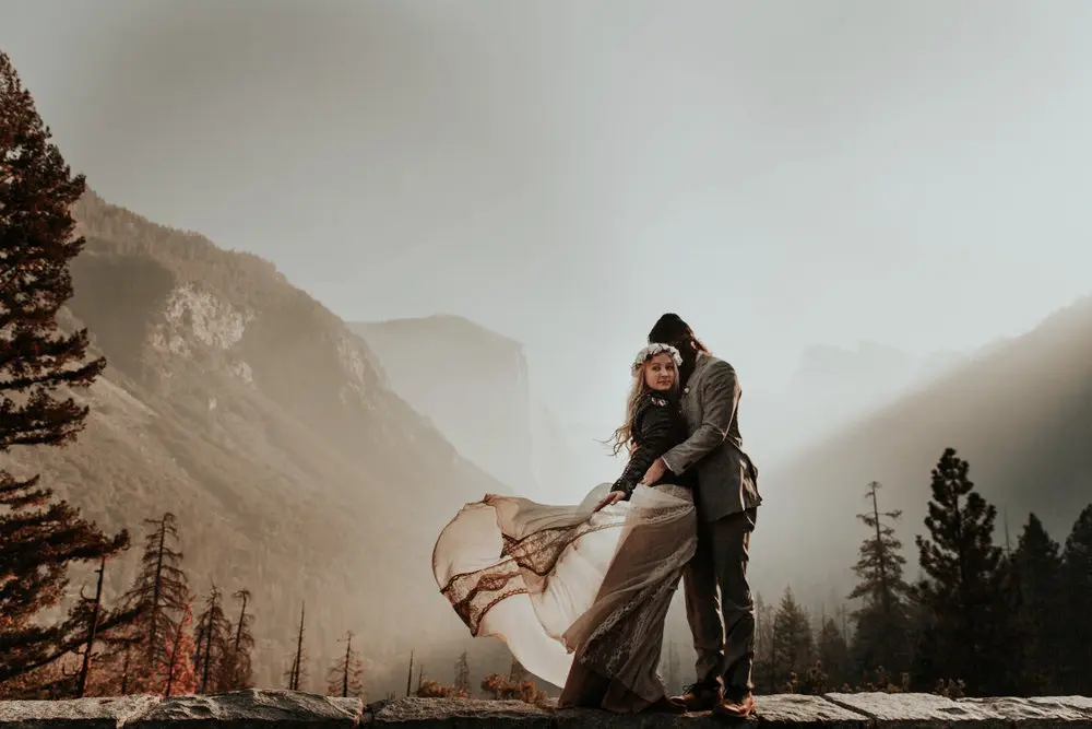 Bride and groom embracing in Yosemite, with the bride's flowing dress caught in the wind and misty mountains behind them.