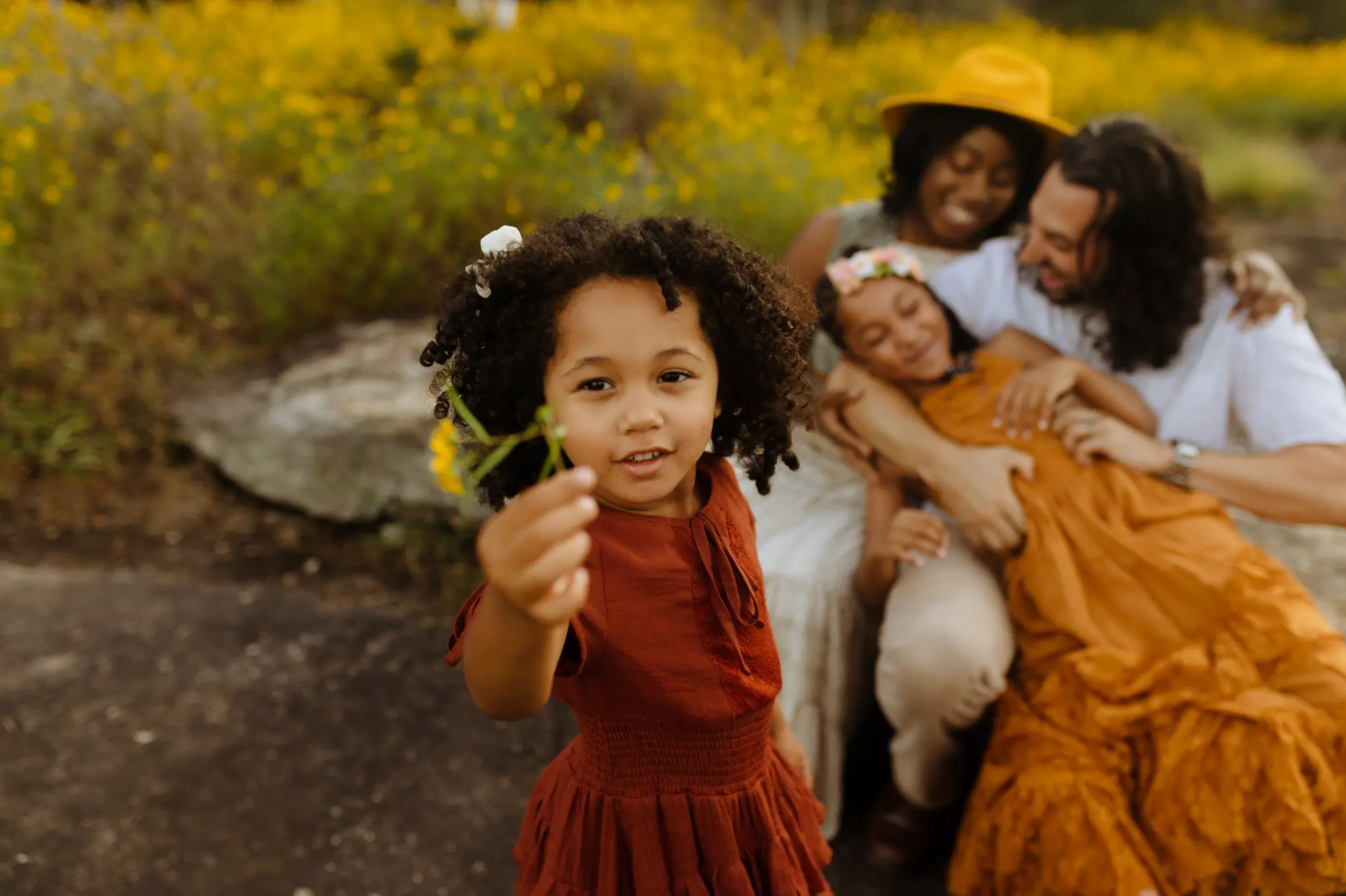 ZION Presets by Meridian enhance this family portrait of a young girl with curly hair holding a flower, with her family lovingly interacting in the background in a field of yellow flowers.