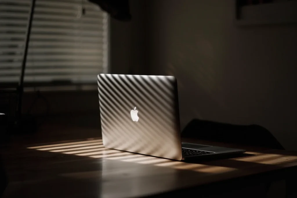 A MacBook Air is sitting on a table lit up by rays of sun through a window shade