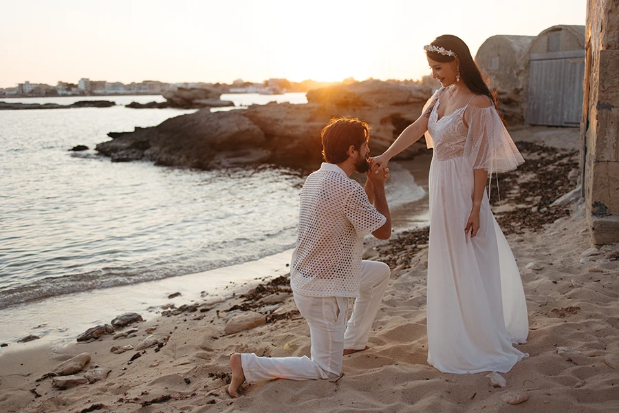 On a sandy beach at sunset, a person kneels before a standing individual who is dressed in a flowing white dress with sheer sleeves and a floral headpiece. The kneeling person is wearing a white, patterned shirt and white pants, holding the hand of the standing individual in a gesture of affection or proposal. The ocean is calm, and the sky is aglow with the golden light of the setting sun, creating a romantic, serene atmosphere. Taken by Mallorca Photographer