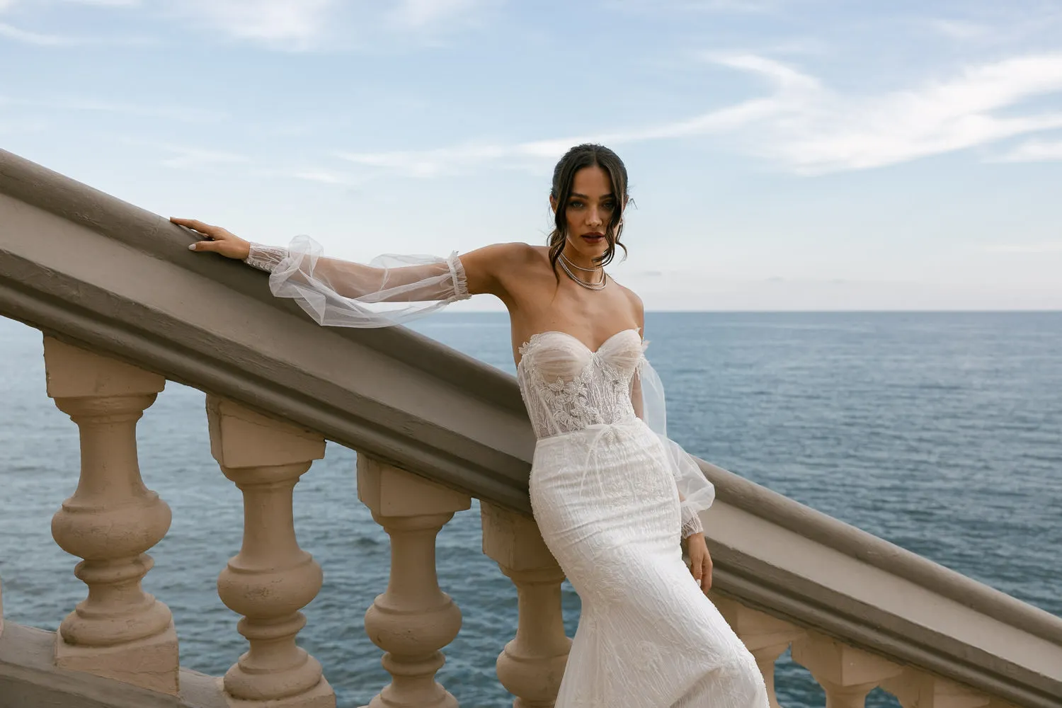 A woman in an off-the-shoulder wedding dress leans against a balustrade overlooking the sea. Her dress features lace detailing and a fitted bodice, and she wears a sheer, tulle sleeve that drapes elegantly. Her dark hair is styled in loose waves, and she accessorizes with a choker necklace. The horizon of the calm sea meets the sky behind her, providing a serene backdrop. 