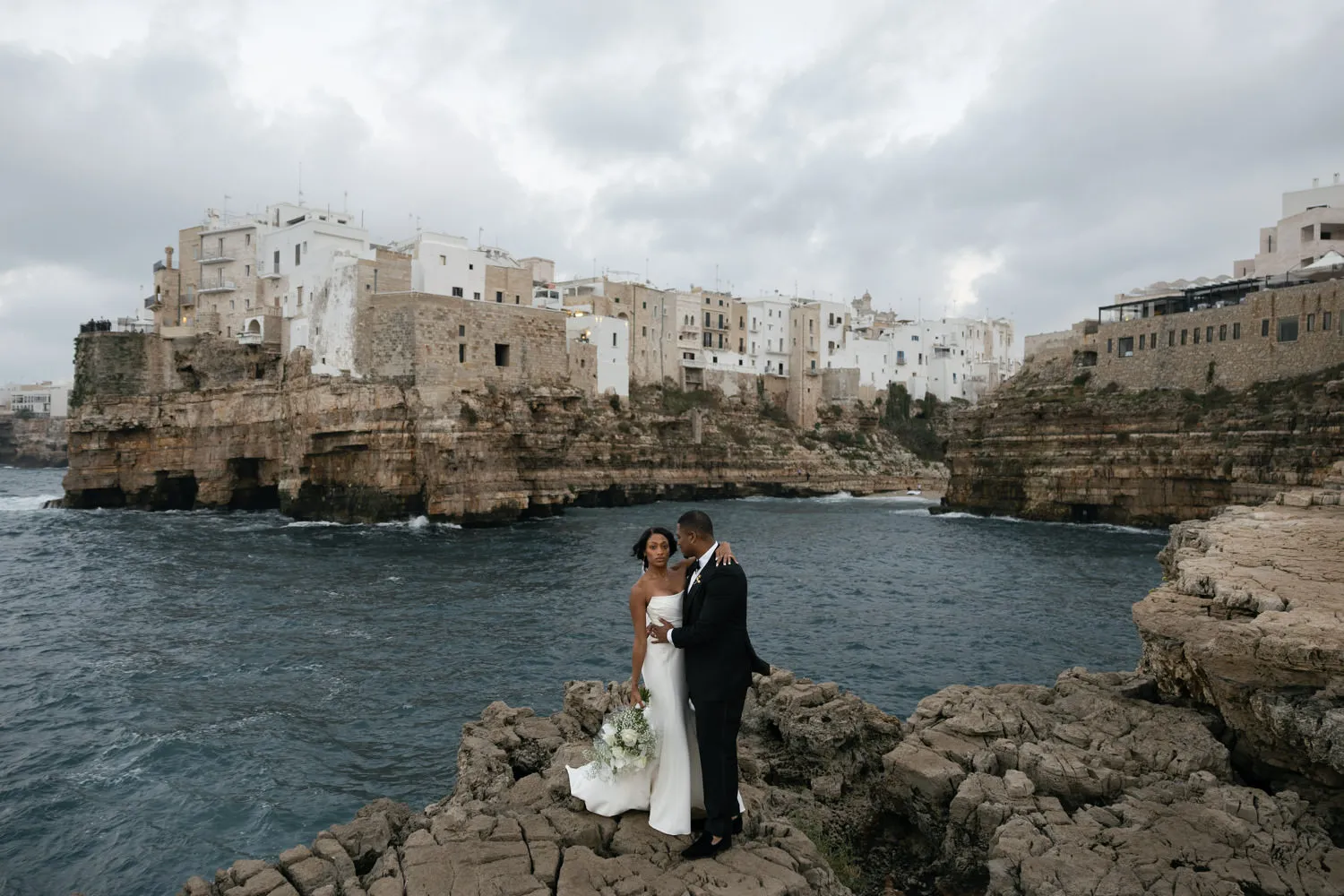A bride and groom are standing on a rocky outcrop by the sea, with the historic white buildings of an old coastal town in the background. The groom, dressed in a black suit, faces the bride, wearing a white strapless wedding gown and holding a bouquet. The sea is dark and choppy, contrasting with the overcast sky, and the town provides a picturesque backdrop to the intimate moment. 
