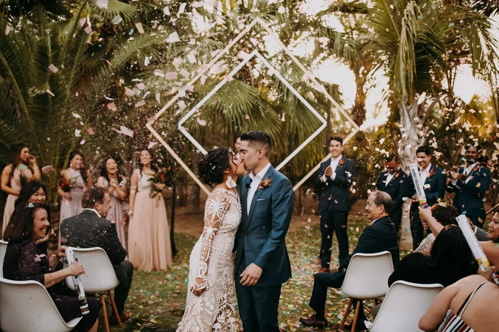 Newlyweds kiss amid confetti at Acre Resort, one of the popular destination wedding venues in Mexico, surrounded by joyous guests and lush palm trees.