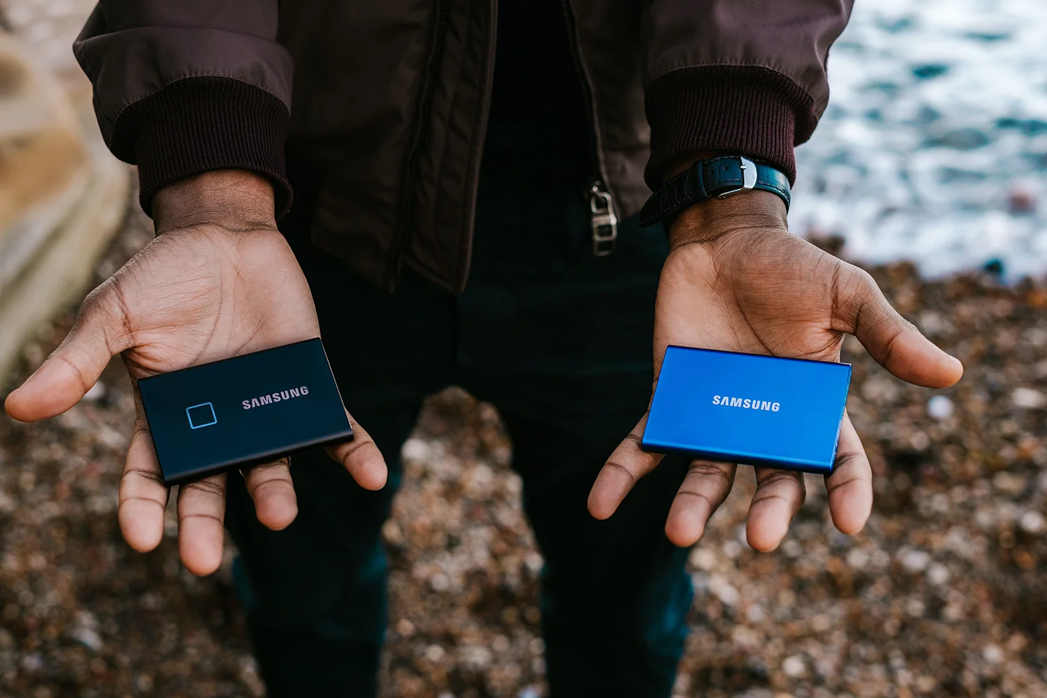 A person holds out two Samsung portable SSDs in each hand, one black and one blue, against a natural outdoor backdrop, symbolizing the intersection of technology and mobility.