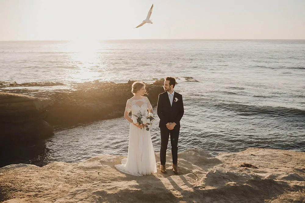 Newlyweds posing by the ocean at sunset with a seagull flying overhead, illustrating a top elopement venue in California.