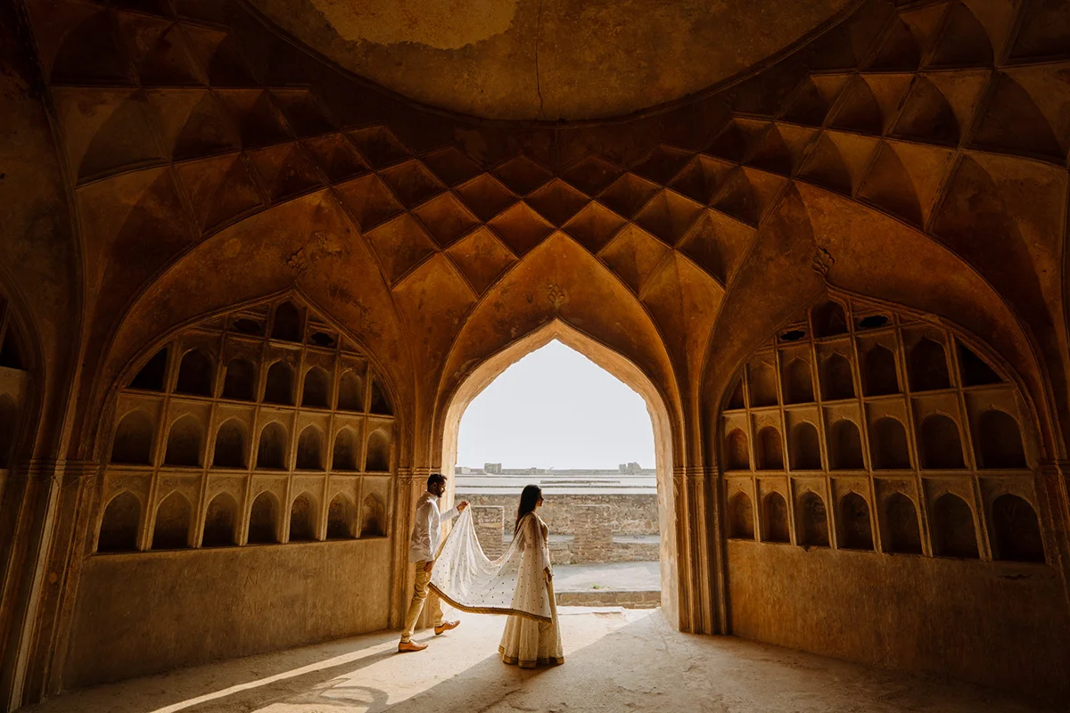 A couple walks hand in hand through an ancient arched hallway, their white traditional attire glowing in the sunlight streaming through, evoking a sense of history and togetherness.