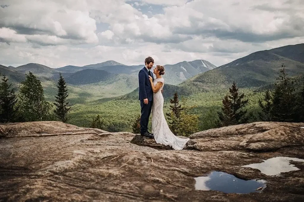 A bride and groom stand together on a rocky cliff with a scenic mountain landscape in the background, captured as part of wedding photography packages. The bride wears a white lace gown, and the groom is in a navy blue suit, both looking lovingly into each other's eyes under a partly cloudy sky.