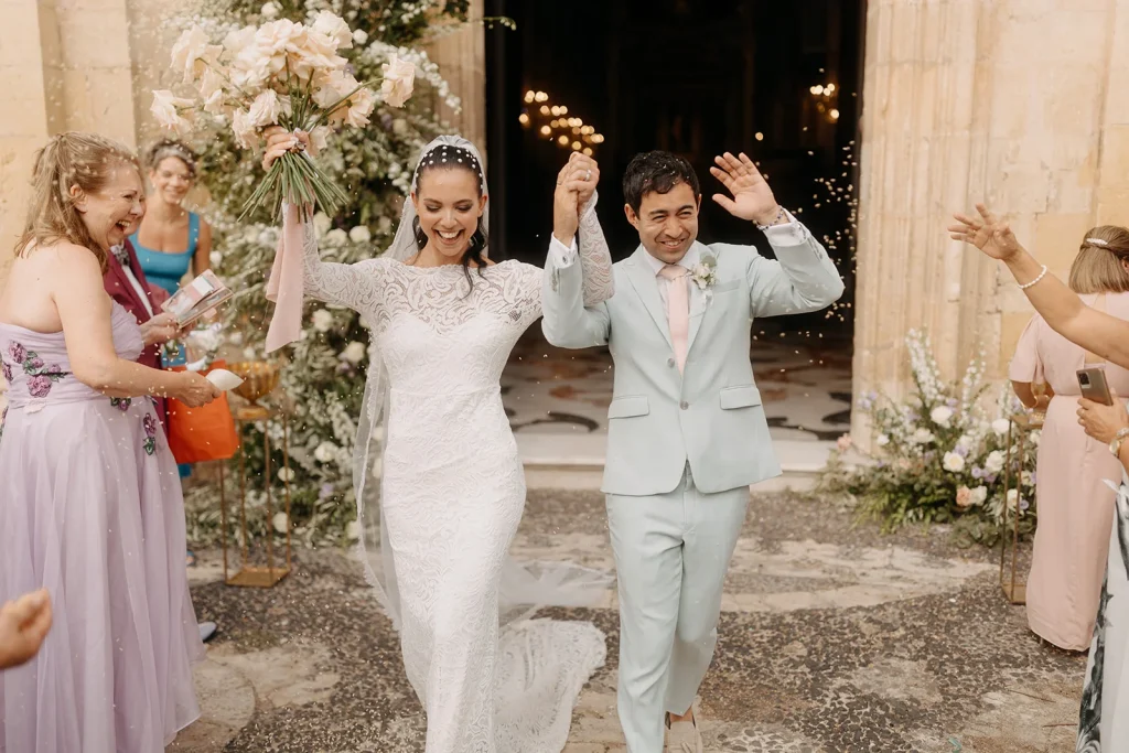 Newlywed couple joyfully exiting an Italian wedding venue, with guests throwing confetti, showcasing the bride's elegant white lace gown and the groom's pastel blue suit.
