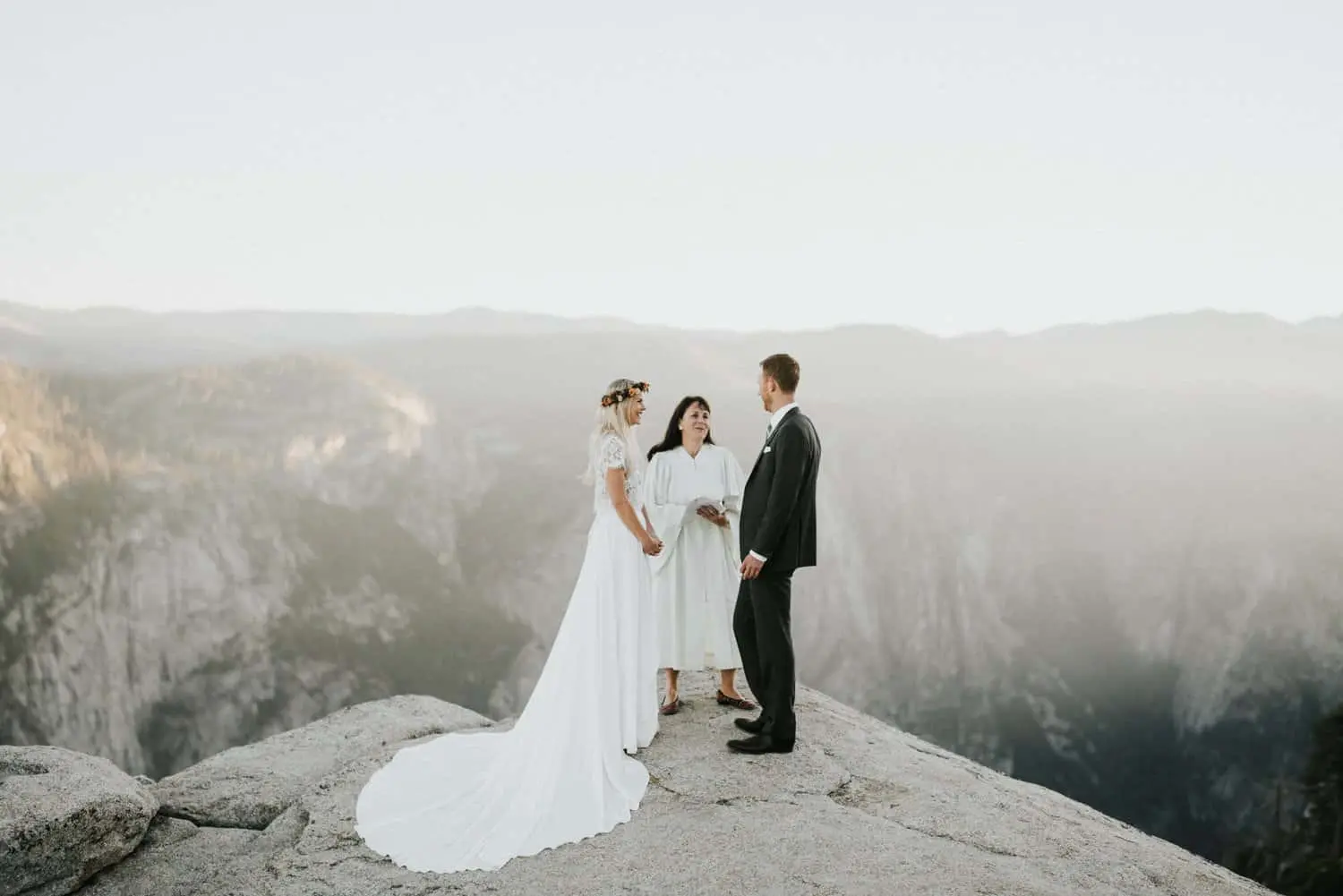 Bride and groom exchanging vows on a cliff during a Yosemite elopement ceremony, with an officiant and misty mountains in the background, after sending out their awesome elopement announcements.